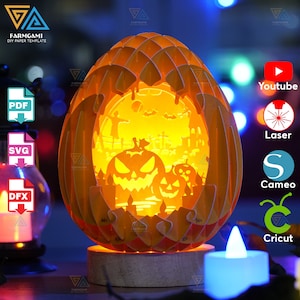 May include: A 3D papercraft pumpkin lamp with a Halloween scene inside. The scene includes a graveyard, a spooky tree, and a cat. The pumpkin is lit from the inside, creating a warm glow. The lamp is made of orange paper and has a wooden base.
