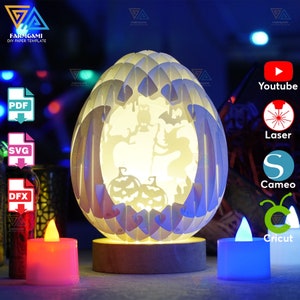 May include: A white papercraft Halloween pumpkin lamp with a spooky design. The lamp is lit from within, casting a warm glow. The lamp is on a wooden base and is surrounded by two tealight candles, one red and one blue.