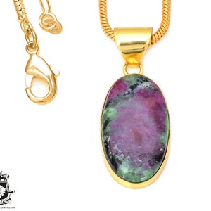 May include: A gold-toned necklace featuring an oval pendant with a ruby zoisite stone. The stone displays a mix of pink, green, and black hues. The necklace has a snake chain and a lobster clasp.