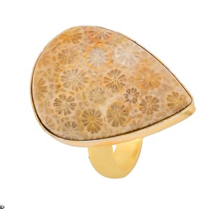 Pode incluir: Um anel de ouro com uma pedra de coral fossilizado em forma de lágrima. A pedra é de cor castanho claro e tem um padrão de pequenas flores brancas.