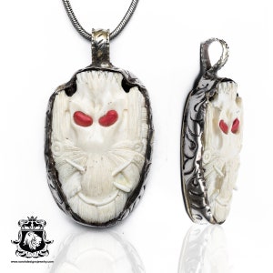 Peut inclure: Un collier pendentif avec un visage sculpté de couleur ivoire et des accents rouges pour les yeux, serti dans un cadre argenté. Le pendentif est présenté de face et de profil, mettant en valeur le design. Le collier est sur une chaîne argentée.