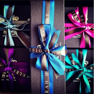 Puede incluir: Cuatro cajas de regalo con tapas negras y lazos de cinta de colores. Las cajas est&aacute;n etiquetadas con el texto "Sarah Design Jewelry".