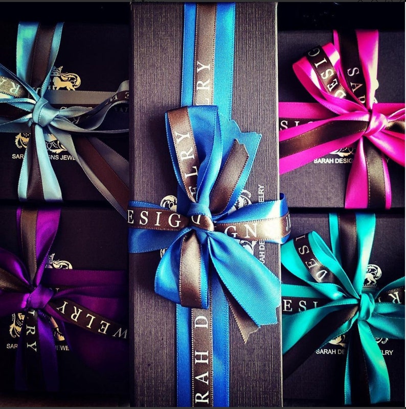 Puede incluir: Cuatro cajas de regalo con tapas negras y lazos de cinta de colores. Las cajas est&aacute;n etiquetadas con el texto "Sarah Design Jewelry".
