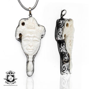 May include: A white bone pendant carved with a detailed face and a crown, set in a silver frame. The pendant is attached to a silver chain.