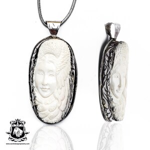 May include: A silver chain necklace with a white carved pendant featuring a woman's face with a feathered headdress. The pendant is oval shaped and has a detailed silver border.