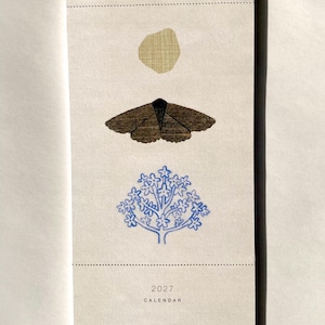 May include: A vertical wall calendar with a white string hanger. The off-white paper features a tan circle, a brown moth illustration, and a blue tree graphic. Below, the text reads 2027 CALENDAR and THIS IS YOUR YEAR.