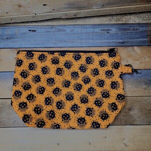 May include: An orange zippered pouch with a repeating pattern of black cat faces. The pouch has a black zipper and a small black loop with a metal clasp. The background is a wooden surface.