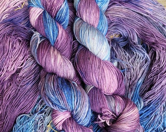 Könnte beinhalten: Zwei Knäuel handgefärbter Wolle in den Farben Lila, Blau und Weiß. Die Wolle ist in einem lockeren Haufen angeordnet, wobei die Enden der Knäuel sichtbar sind.