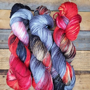May include: Three skeins of hand-dyed yarn in shades of red, black, and gray. The yarn is arranged in a criss-cross pattern.