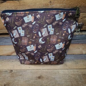 May include: A dark brown zippered pouch featuring a repeating pattern of white and tan illustrations including tarot cards, crescent moons, candles, crystals, keys, mortar and pestle sets, and small floral sprigs. The pouch has a black zipper.