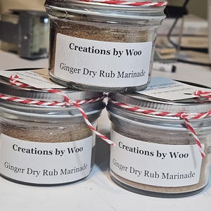 May include: Three glass jars with red and white twine tied around the lids. Each jar contains a brown spice blend and has a white label that reads "Creations by Woo Ginger Dry Rub Marinade".