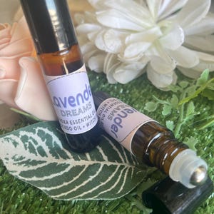 May include: Two amber glass roller bottles with black caps. The bottles are labeled "Lavender Dreams" and contain essential oils. The bottles are sitting on a green surface.