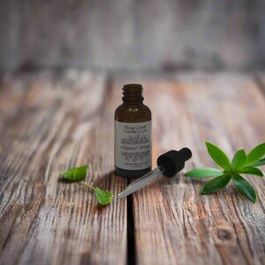 May include: A brown glass bottle with a white label that reads "Rose Creek Candle Co." and "Magical Dream." A black dropper and green leaves are next to the bottle on a wooden surface.