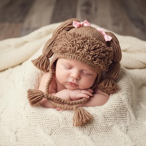 May include: A brown crocheted hat featuring a textured loop-stitch brim, two long braided pigtails with tassel ends, and two small light pink satin bows attached to the top. The hat is designed with ear flaps that tie under the chin.