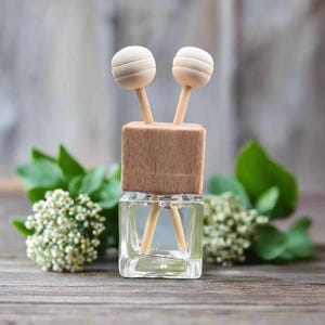 May include: A clear glass aromatherapy diffuser bottle with a wooden top and two wooden sticks topped with round, light-colored wooden balls. The diffuser sits on a wooden surface, surrounded by green leaves and white flowers.
