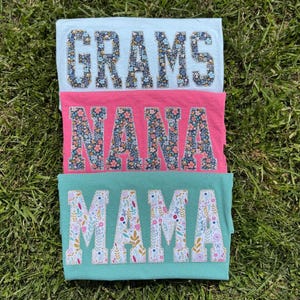 Pu&ograve; includere: Tre magliette piegate in azzurro, rosa e verde acqua, ognuna con una parola a fantasia floreale: "GRAMS", "NANA" e "MAMA". Le magliette sono disposte su erba verde, creando un regalo colorato e personalizzato.