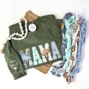 May include: Green sweatshirt with the word "MAMA" in large, colorful letters. The letters are made of fabric with different animal and sports-themed patterns. The sweatshirt has a small embroidered heart and the names "Luka" and "Henri" on the sleeve.