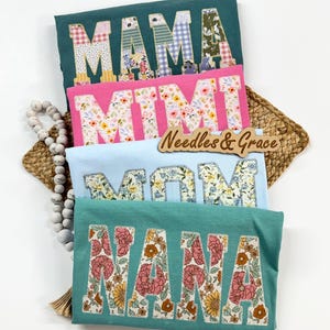 Pu&ograve; includere: Quattro magliette in verde acqua, rosa e azzurro, ognuna con un nome diverso in lettere patchwork floreali: MAMA, MIMI, MOM e NANA. &Egrave; visibile anche un cartello di legno con la scritta "Needles & Grace".