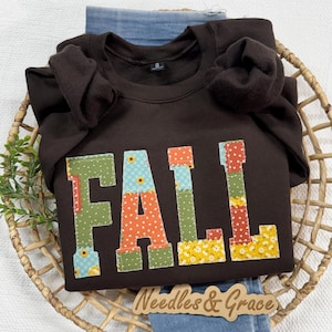 May include: A dark brown crewneck sweatshirt featuring the word FALL in large, block-style letters. Each letter is filled with a different colorful fabric pattern, including green, orange, and yellow floral and polka dot prints on a dark background.