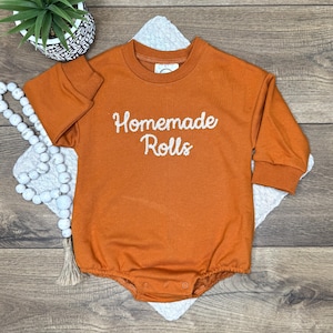 May include: An orange long-sleeved baby romper with the words "Homemade Rolls" in white script. The romper has a round neckline and snap closures at the bottom. A small potted plant and decorative beads are in the background.