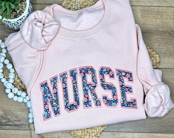 May include: A light pink sweatshirt with the word "NURSE" in large, floral-patterned letters across the chest. The sweatshirt is folded on a woven mat, with a small potted plant and decorative beads nearby.