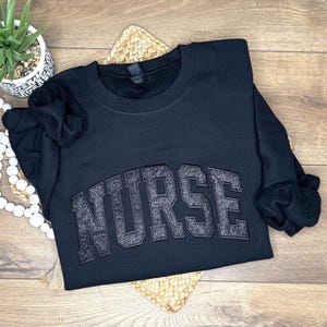 May include: Black crewneck sweatshirt with the word "NURSE" in a glittery, block letter design across the chest. The sweatshirt is displayed on a wooden surface with a small potted plant and decorative beads.