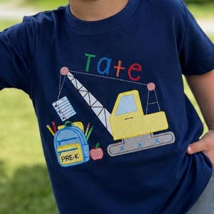 May include: A navy blue short-sleeved t-shirt featuring an embroidered yellow construction crane, a blue backpack with the text PRE-K, a red apple, and a notebook. The name Tate is stitched in multicolored letters above the crane design.