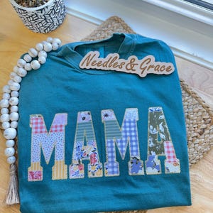 Pu&ograve; includere: T-shirt color verde acqua con la scritta "MAMA" applicata in vari tessuti fantasia. La maglietta &egrave; esposta su una superficie di legno con un tappetino intrecciato e perline decorative. Un cartello di legno con la scritta "Needles & Grace" &egrave; sopra la maglietta.