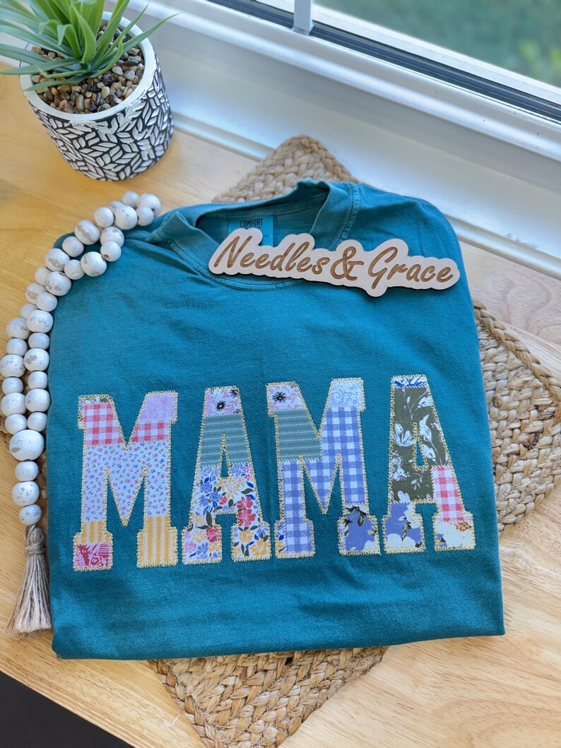 Pu&ograve; includere: T-shirt color verde acqua con la scritta "MAMA" applicata in vari tessuti fantasia. La maglietta &egrave; esposta su una superficie di legno con un tappetino intrecciato e perline decorative. Un cartello di legno con la scritta "Needles & Grace" &egrave; sopra la maglietta.