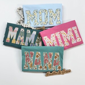 Pu&ograve; includere: Cinque magliette colorate con lettere floreali che compongono "MOM", "MAMA", "MIMI" e "NANA". Le magliette sono in azzurro, verde acqua e rosa. Un cartello di legno con la scritta "Needles & Grace" &egrave; in basso.