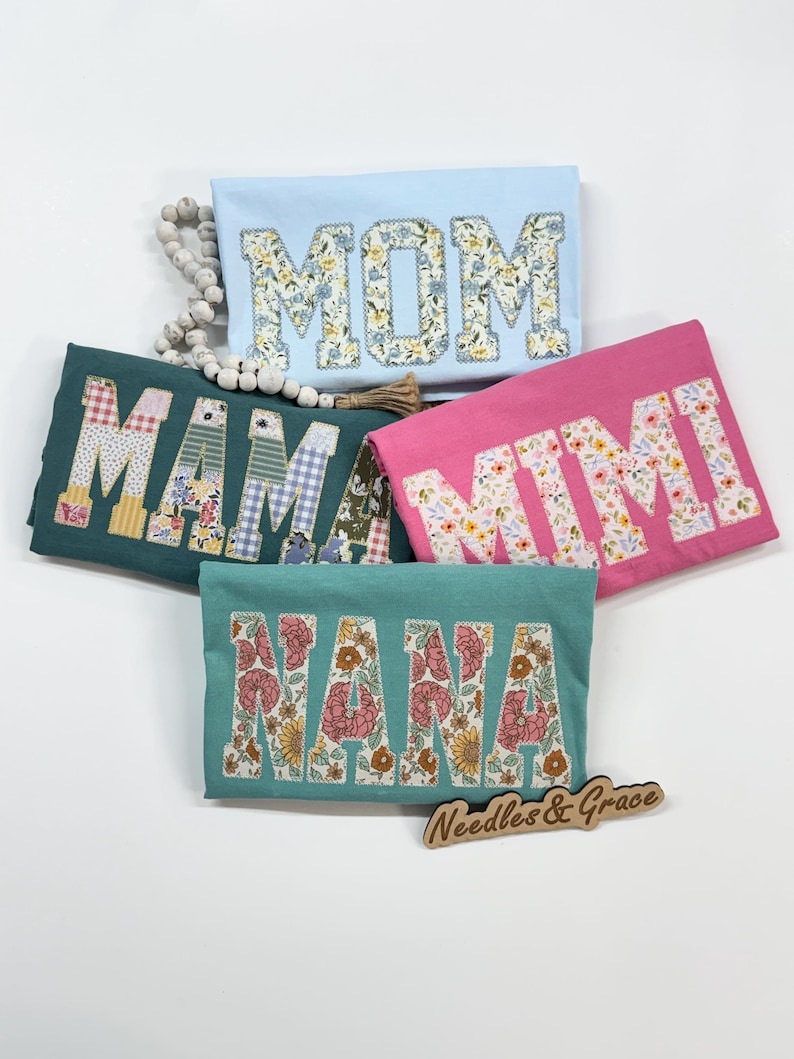 Pu&ograve; includere: Cinque magliette colorate con lettere floreali che compongono "MOM", "MAMA", "MIMI" e "NANA". Le magliette sono in azzurro, verde acqua e rosa. Un cartello di legno con la scritta "Needles & Grace" &egrave; in basso.