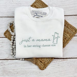 May include: White sweatshirt with the text "just a mama in her string cheese era" in a script font. The sweatshirt has a small graphic of a string cheese stick. The brand logo "Needles & Grace" is at the top.