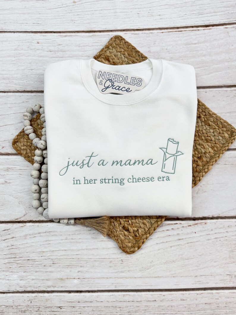 May include: White sweatshirt with the text "just a mama in her string cheese era" in a script font. The sweatshirt has a small graphic of a string cheese stick. The brand logo "Needles & Grace" is at the top.
