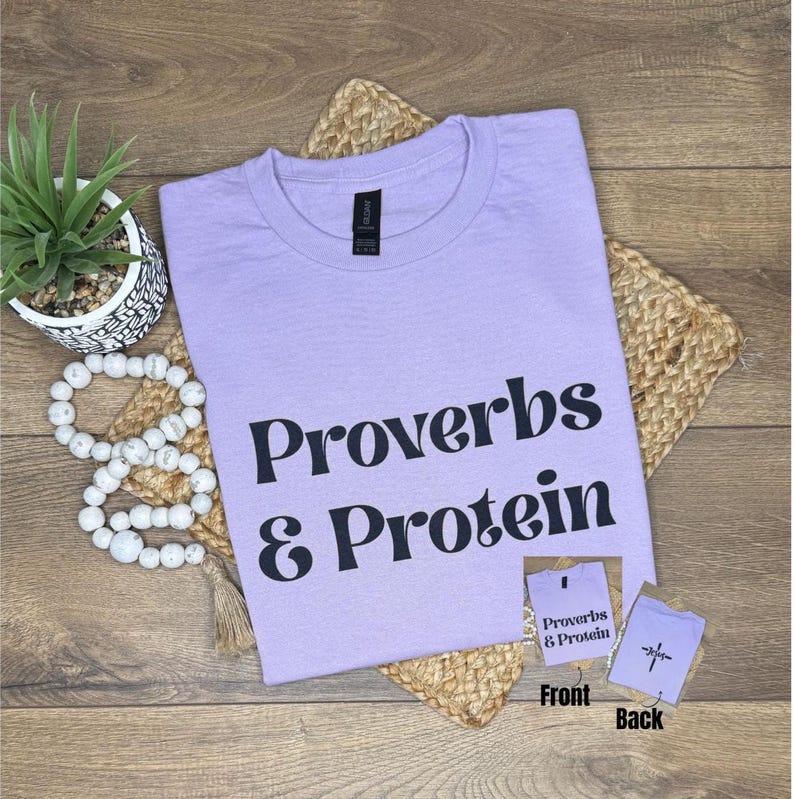 May include: A lilac t-shirt with the text "Proverbs & Protein" in black lettering. The shirt is placed on a woven mat, alongside a small potted plant and a string of white beads. A small card shows the front and back of the shirt.
