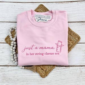 May include: A light pink sweatshirt with the text "just a mama in her string cheese era" in a cursive font. A string cheese graphic is also printed on the front. The sweatshirt has a crew neck and long sleeves.