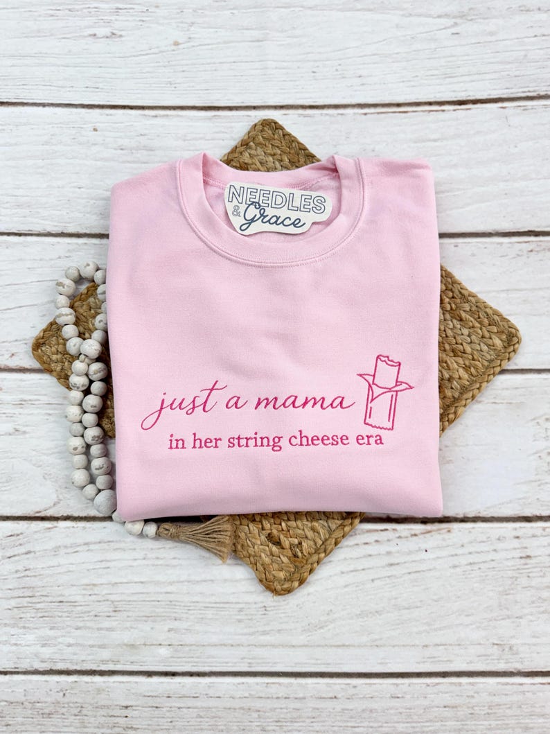 May include: A light pink sweatshirt with the text "just a mama in her string cheese era" in a cursive font. A string cheese graphic is also printed on the front. The sweatshirt has a crew neck and long sleeves.