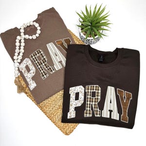 May include: Two brown sweatshirts with the word "PRAY" appliqued on the front. The letters are made of various patterned fabrics, including floral, plaid, and striped designs. A white beaded necklace and a small potted plant are also in the image.