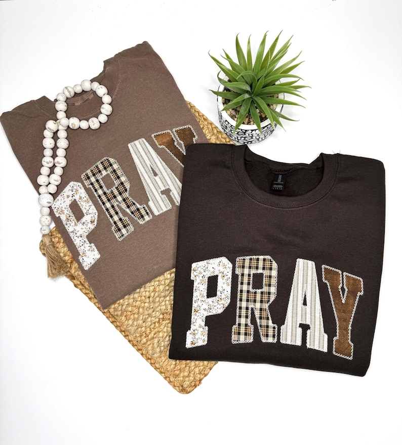 May include: Two brown sweatshirts with the word "PRAY" appliqued on the front. The letters are made of various patterned fabrics, including floral, plaid, and striped designs. A white beaded necklace and a small potted plant are also in the image.