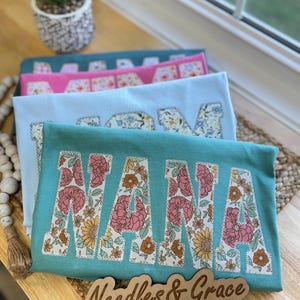 Pu&ograve; includere: Tre magliette sovrapposte, turchese, rosa e azzurro chiaro, ognuna con la scritta "NANA" in lettere floreali. Su una superficie di legno, un cartello di legno recita "Needles & Grace".