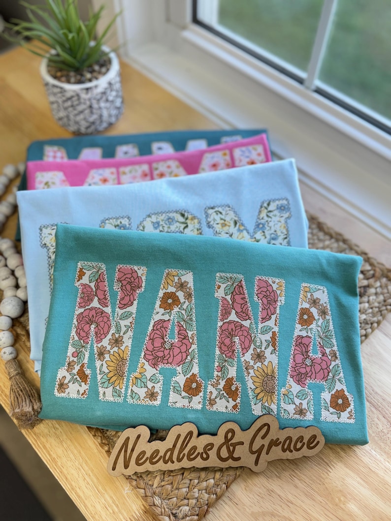Pu&ograve; includere: Tre magliette sovrapposte, turchese, rosa e azzurro chiaro, ognuna con la scritta "NANA" in lettere floreali. Su una superficie di legno, un cartello di legno recita "Needles & Grace".