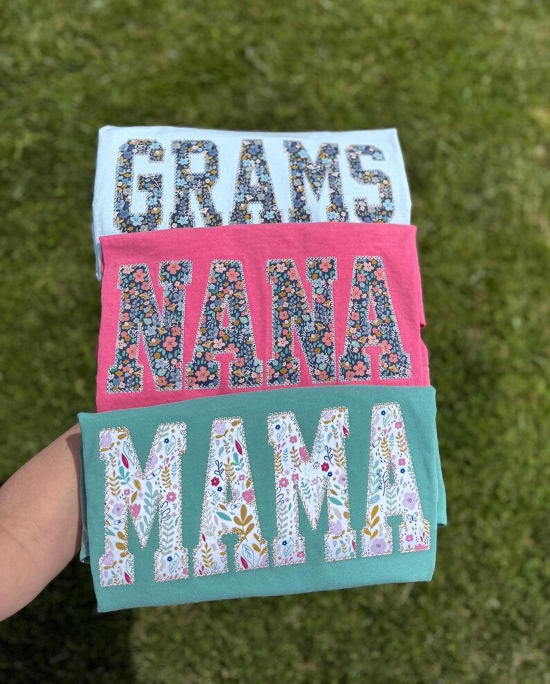 Pu&ograve; includere: Tre magliette colorate sovrapposte. La maglietta superiore &egrave; azzurra con la scritta "GRAMS" in stampa floreale. La maglietta centrale &egrave; rosa con "NANA" in stampa floreale. La maglietta inferiore &egrave; verde acqua con "MAMA" in stampa floreale.