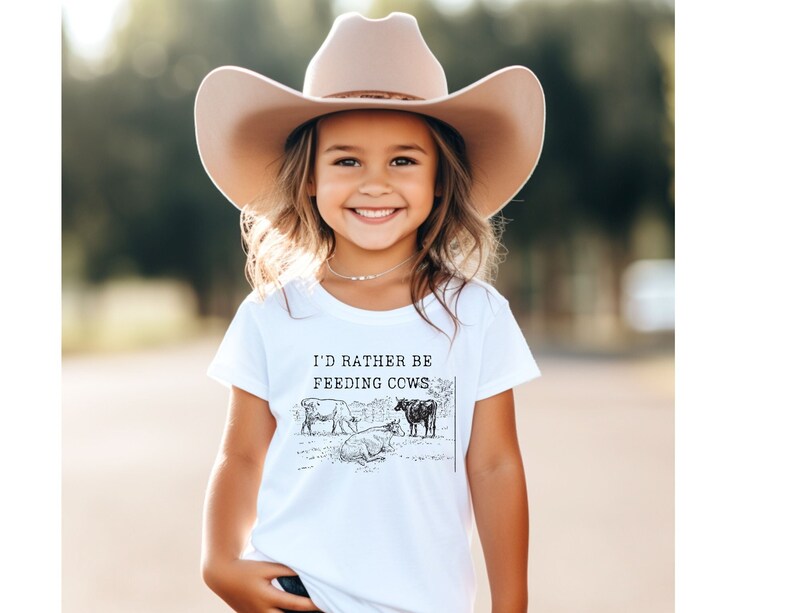 Op de afbeelding: Een wit T-shirt met een zwart-witte illustratie van koeien en de tekst "I'd rather be feeding cows". Een jong meisje draagt het shirt en een bruine cowboyhoed.