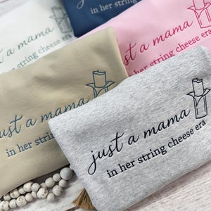 May include: A collection of folded sweatshirts in various colors, including gray, pink, tan, and navy blue. Each sweatshirt features the embroidered phrase "just a mama in her string cheese era" and a string cheese graphic.