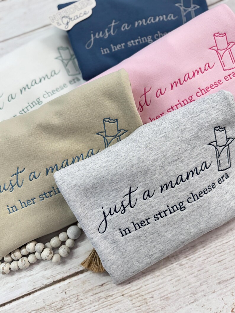 May include: A collection of folded sweatshirts in various colors, including gray, pink, tan, and navy blue. Each sweatshirt features the embroidered phrase "just a mama in her string cheese era" and a string cheese graphic.