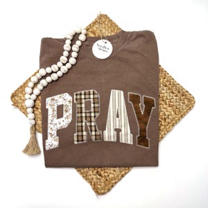 May include: A brown sweatshirt with the word "PRAY" appliqued across the front in various patterns. The letters are floral, plaid, striped, and corduroy. A beaded garland and a tag that says "Needles Grace" are also in the image.
