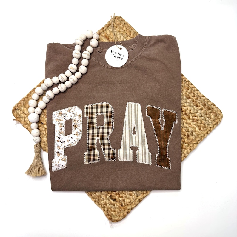 May include: A brown sweatshirt with the word "PRAY" appliqued across the front in various patterns. The letters are floral, plaid, striped, and corduroy. A beaded garland and a tag that says "Needles Grace" are also in the image.