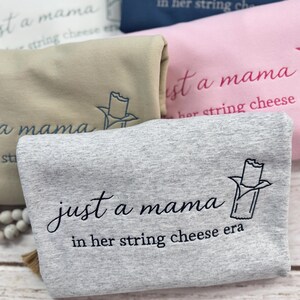 May include: Close-up of several folded sweatshirts in various colors, including gray, pink, and navy blue. Each sweatshirt features the embroidered text "just a mama in her string cheese era" and a string cheese graphic.
