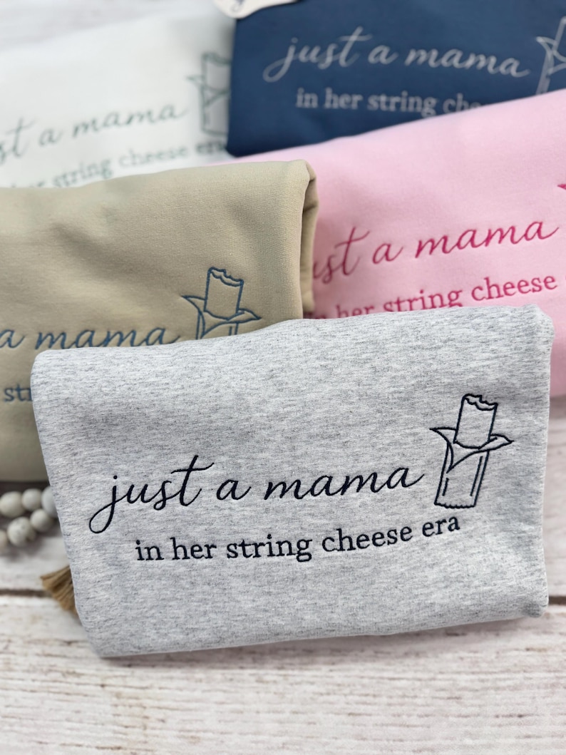 May include: Close-up of several folded sweatshirts in various colors, including gray, pink, and navy blue. Each sweatshirt features the embroidered text "just a mama in her string cheese era" and a string cheese graphic.