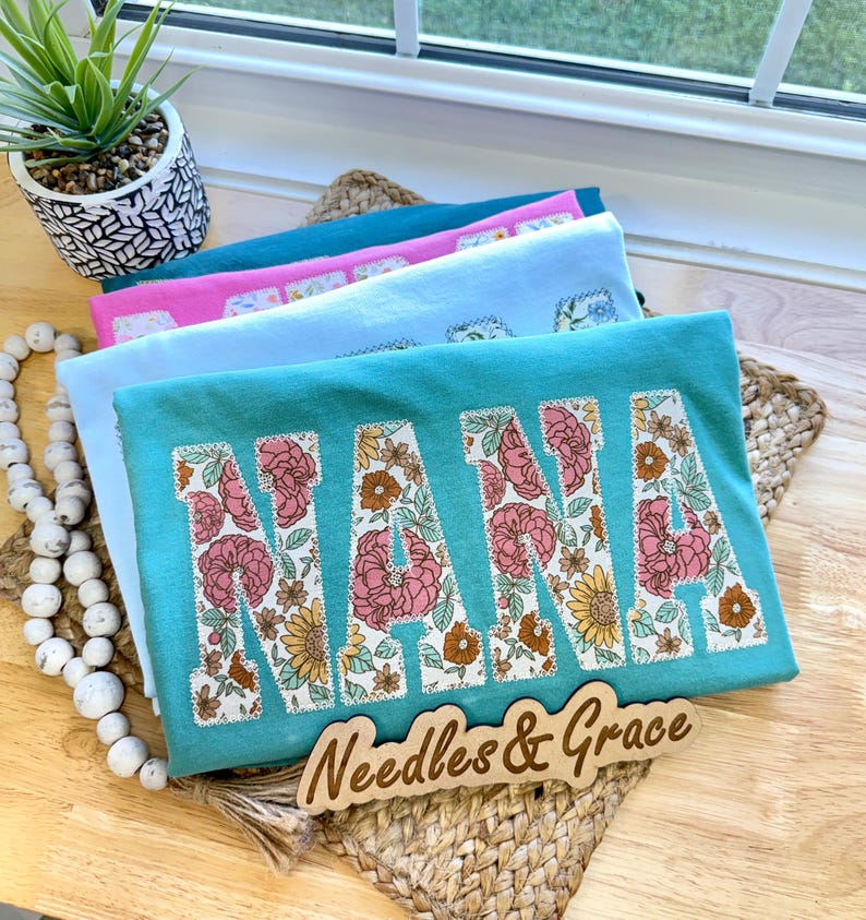Pu&ograve; includere: Una pila di magliette colorate con la parola "NANA" in grandi lettere floreali. Le magliette sono turchese, rosa e azzurro. Un cartello di legno con la scritta "Needles & Grace" &egrave; davanti alle magliette.