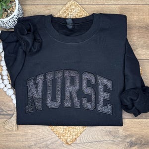 May include: A black crewneck sweatshirt with the word "NURSE" in large, glittery, gray block letters across the chest. The sweatshirt is folded on a woven mat, with a small potted plant and a string of white beads in the background.